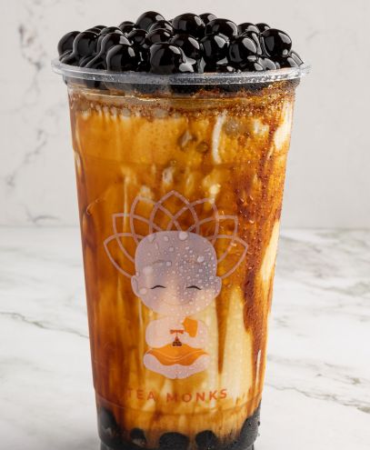 A clear plastic cup filled with brown sugar bubble tea, featuring swirls of milk and brown sugar syrup, topped with glossy black tapioca pearls, set against a light marble background. The cup has a cartoon monk logo printed on it.